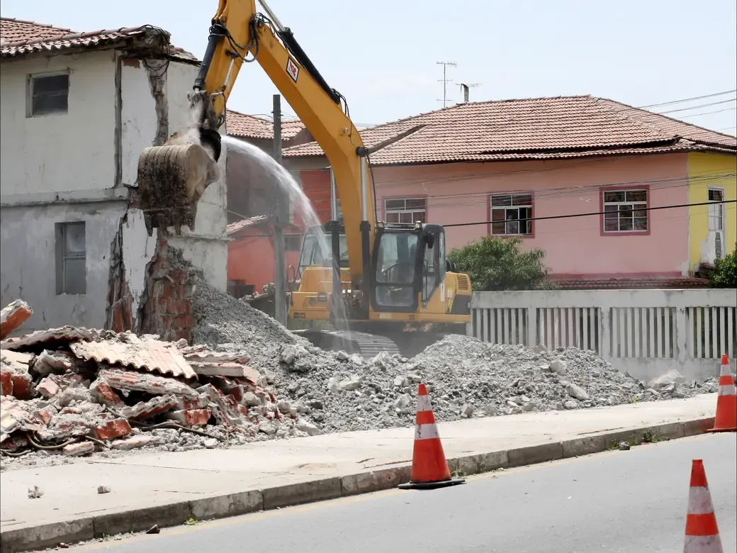 Desmonte de edificação antiga com escavadeira, com aspersão de água para conter a poeira e a via isolada.
